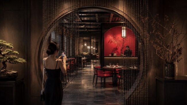 Guest photographing a moon gate entrance with bead curtains, dark wood, and red interior accents in a Chinese-inspired restaurant.