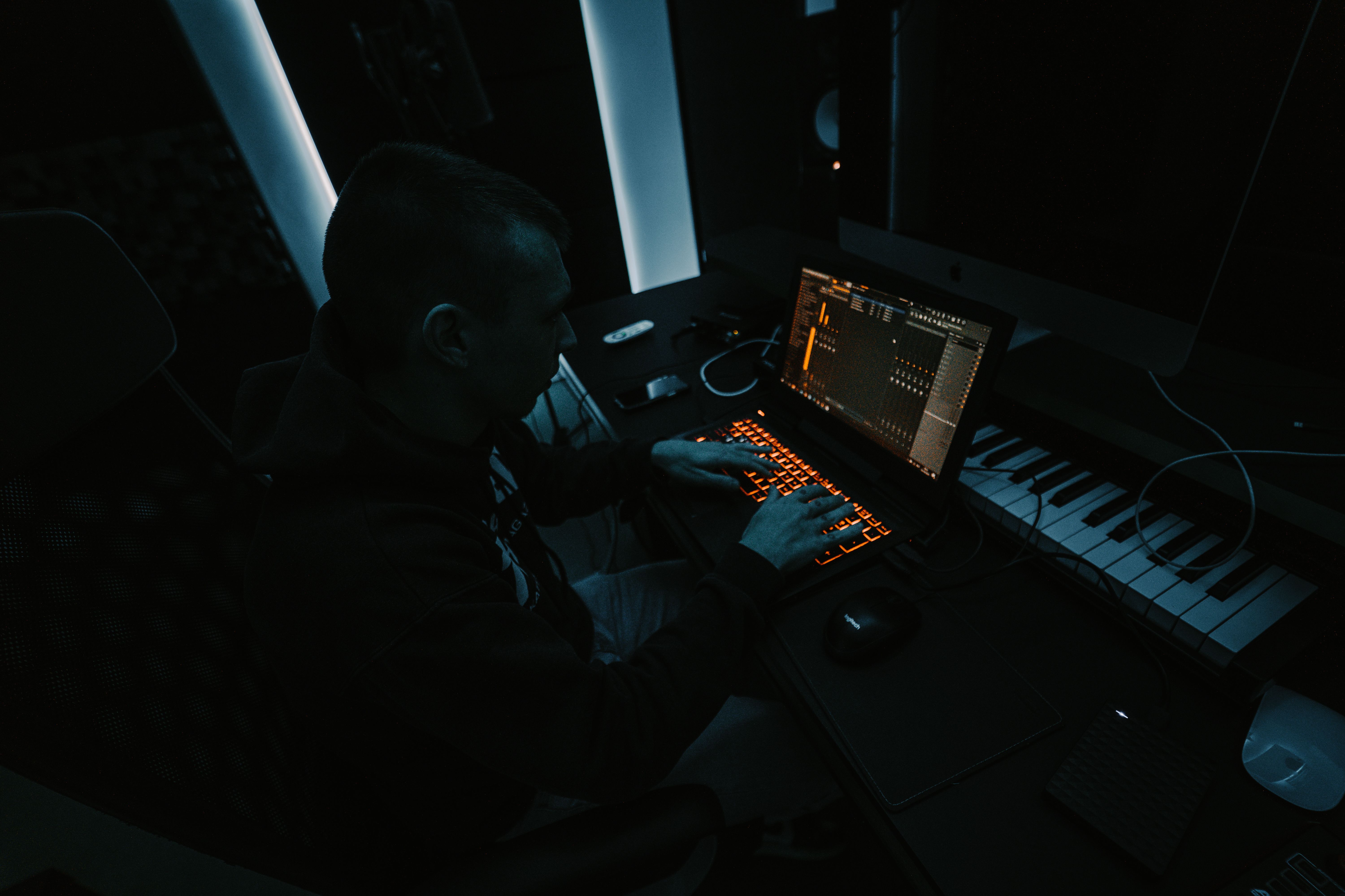 Producer composing music at a keyboard and laptop in a softly lit studio.